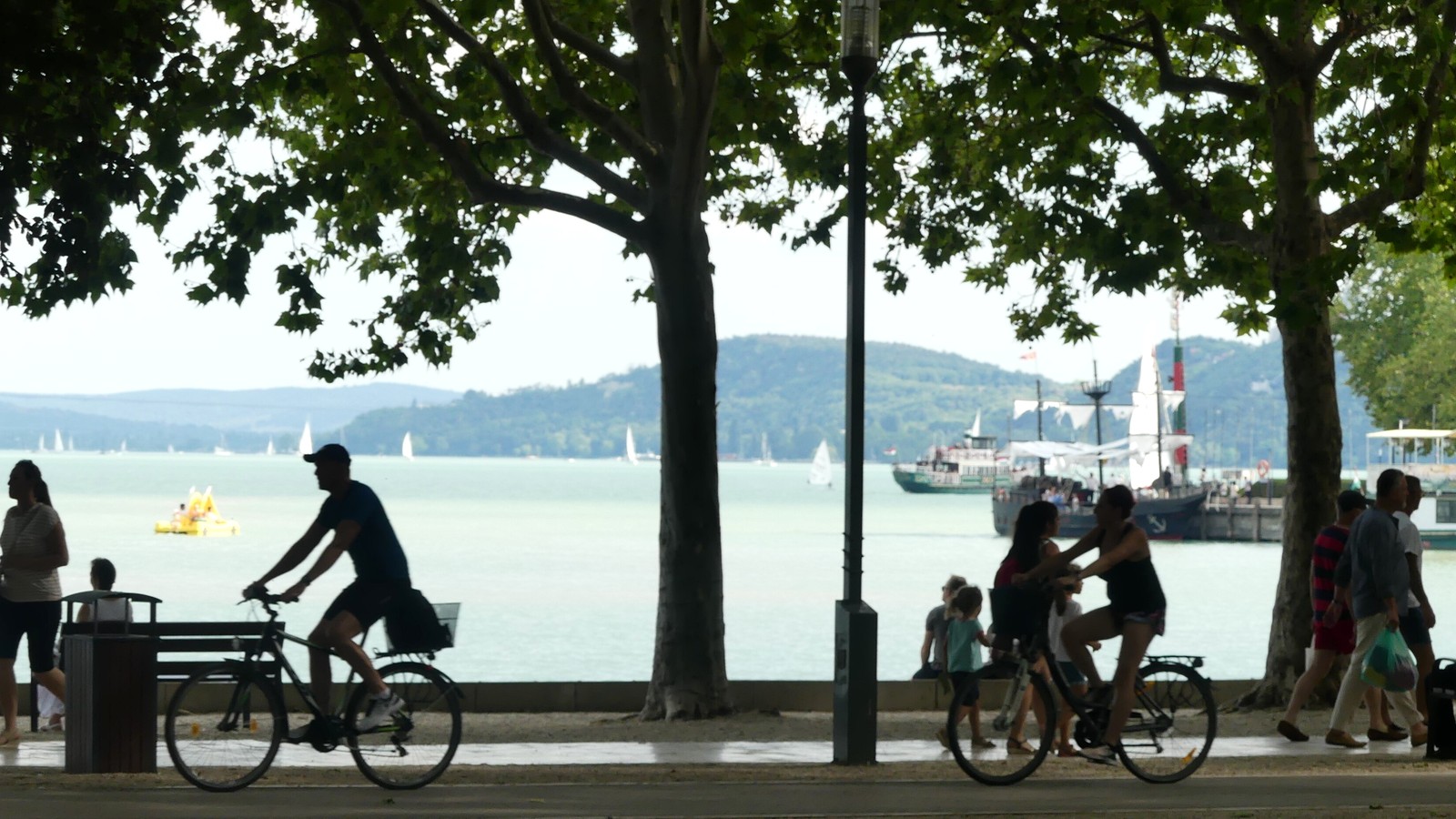 Balatonfüred, a Tagore sétány platánfái alatt kerékpározók és sétálók, mögöttük a Balaton vitorlásokkal és a tihanyi félsziget dombjaival.