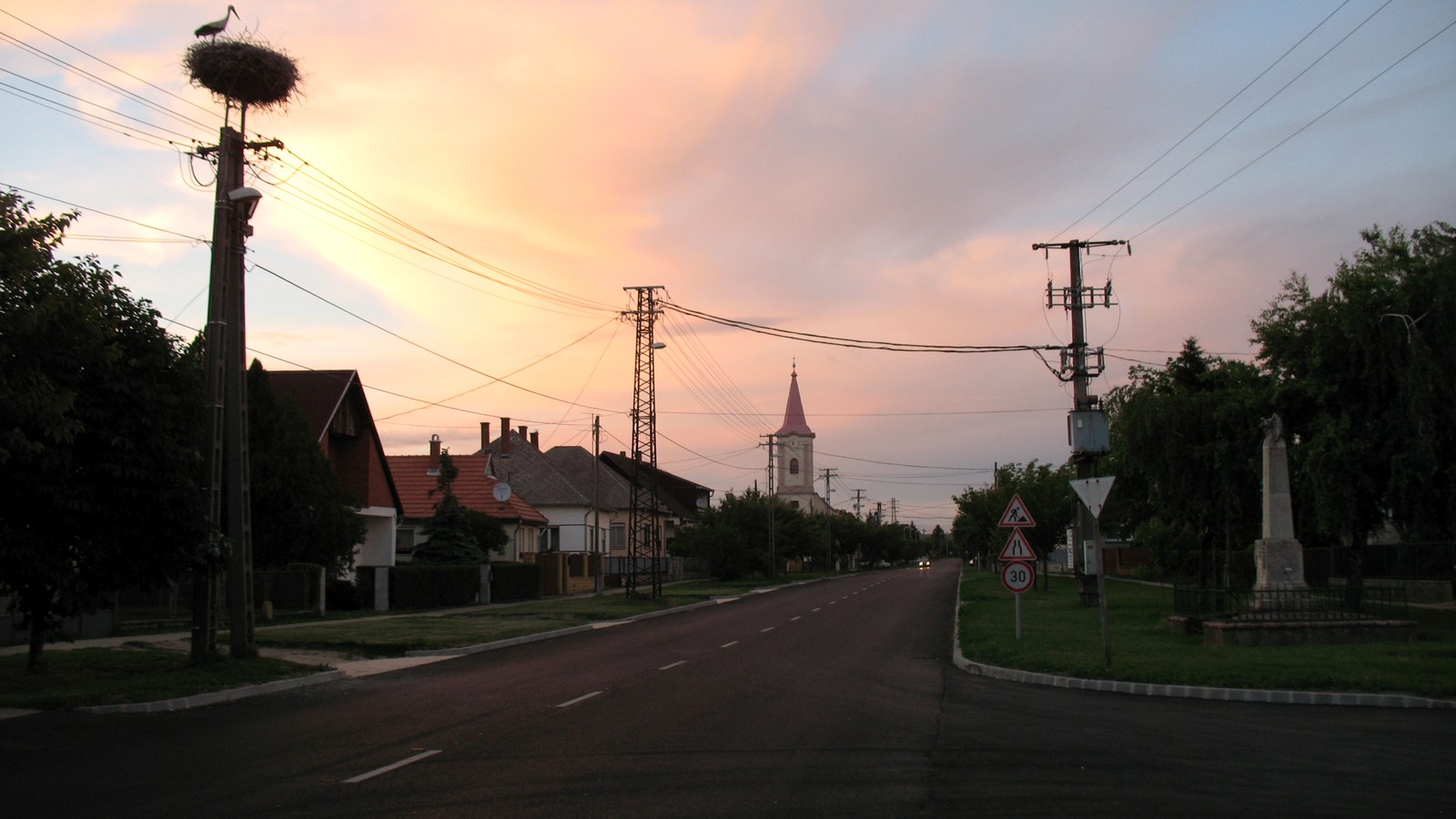 Bana főutcája alkonyatkor, egy oszlop tetején gólyafészekkel, háttérben a templom tornyával.