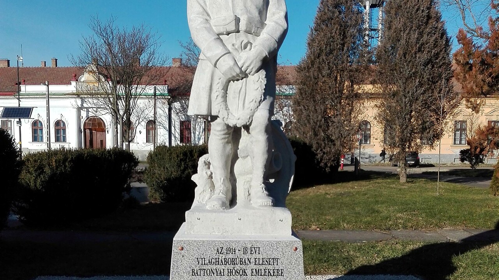 Battonya, az első világháborús hősi emlékmű fehér kőszobra feliratos talapzaton, mögötte emeletes középületek és fák.