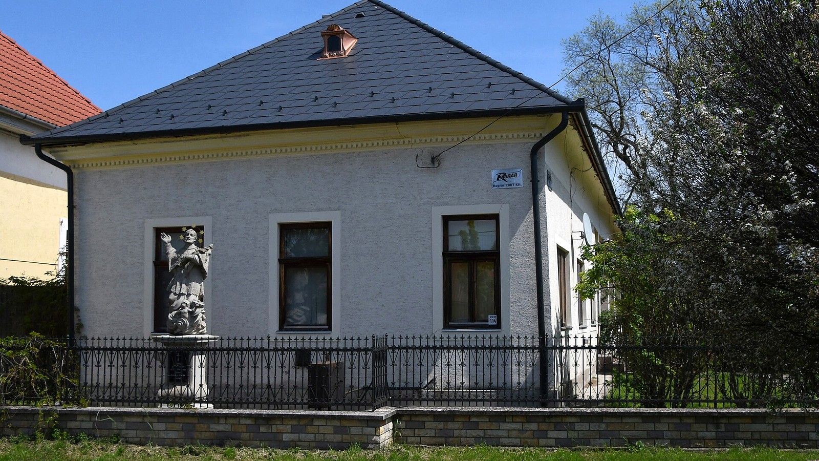 Besenyőtelek utcaképe egy régi, kerítéssel övezett épülettel, homlokzati fülkéjében Nepomuki Szent János szobrával.