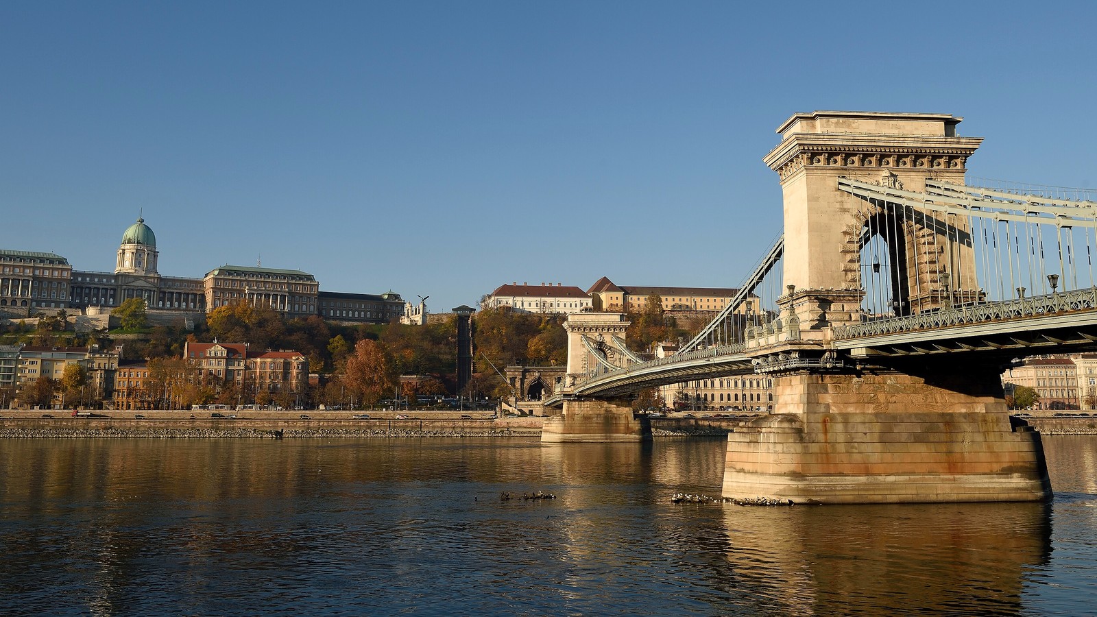 Budapest, a Budavári Palota kupolás épületsora a Várhegyen és a Széchenyi Lánchíd kőpillére a Duna fölött, derült ég alatt.