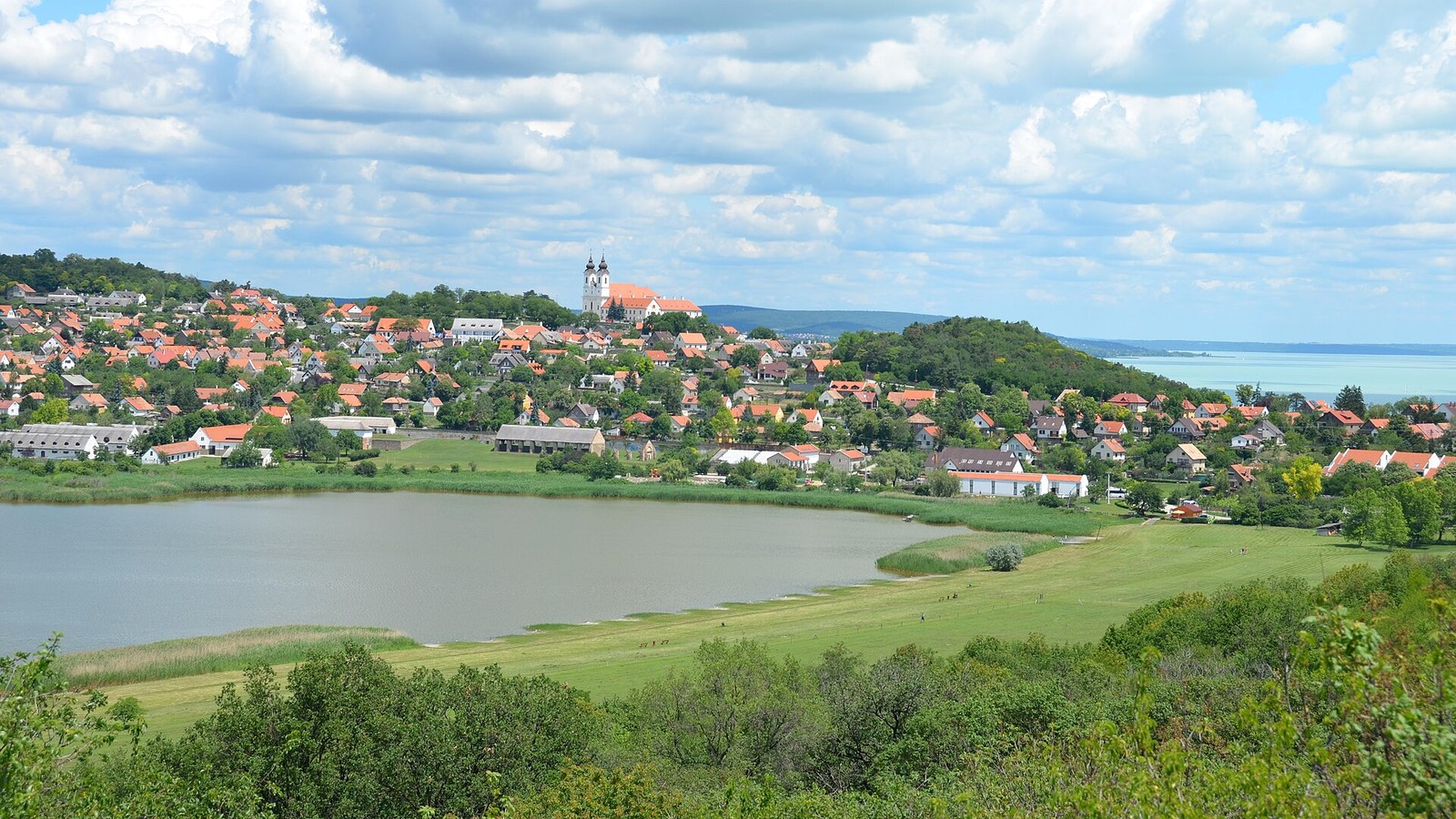 Tihany, a Belső-tó és a partját övező rét, mögötte a domboldalon piros tetős házakkal, a távolban az apátság tornyaival és a Balatonnal.