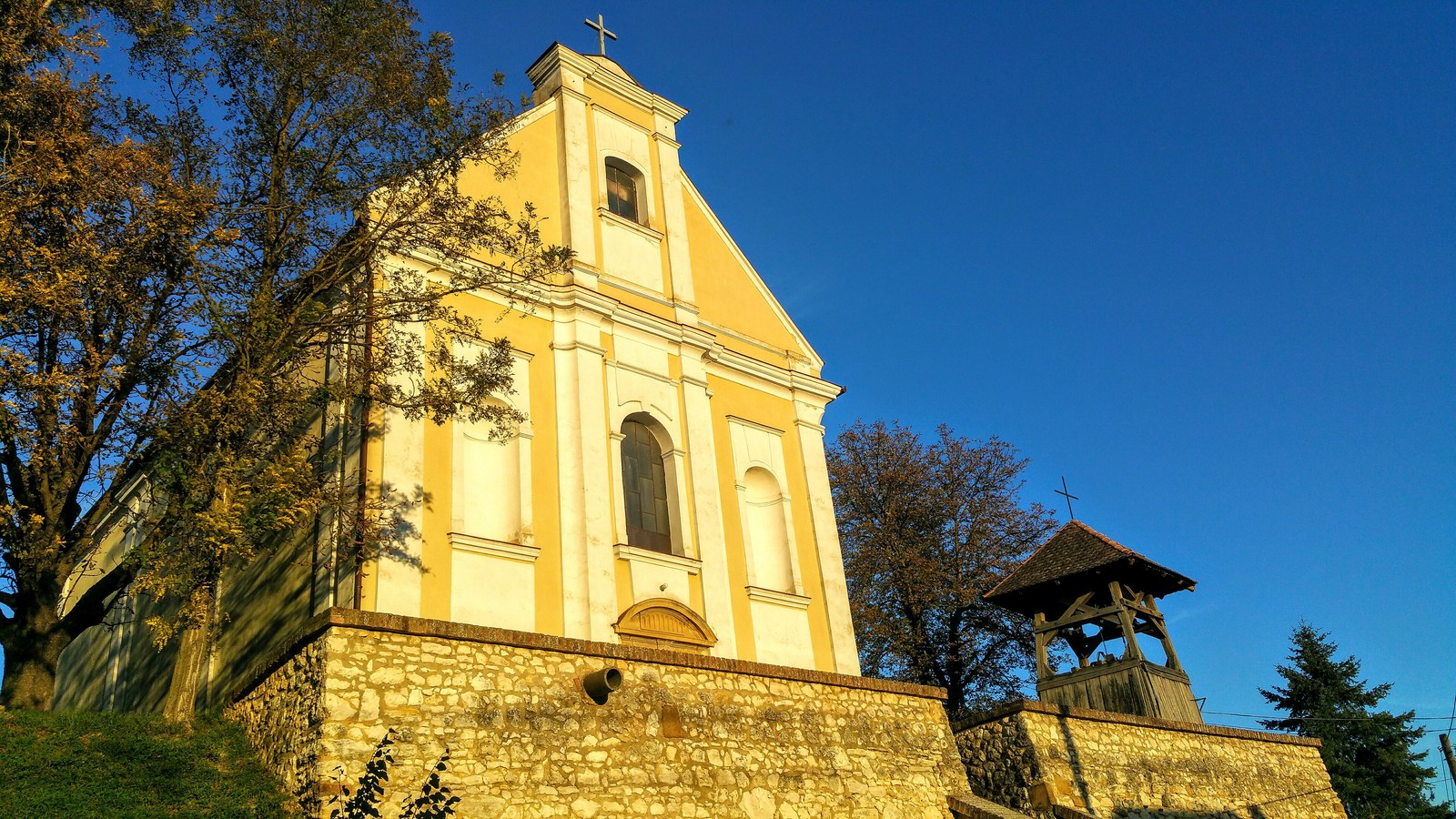 Erzsébet, a Szent Erzsébet-templom sárga homlokzata a kőfal fölött, mellette a fa harangláb, őszi fák között.
