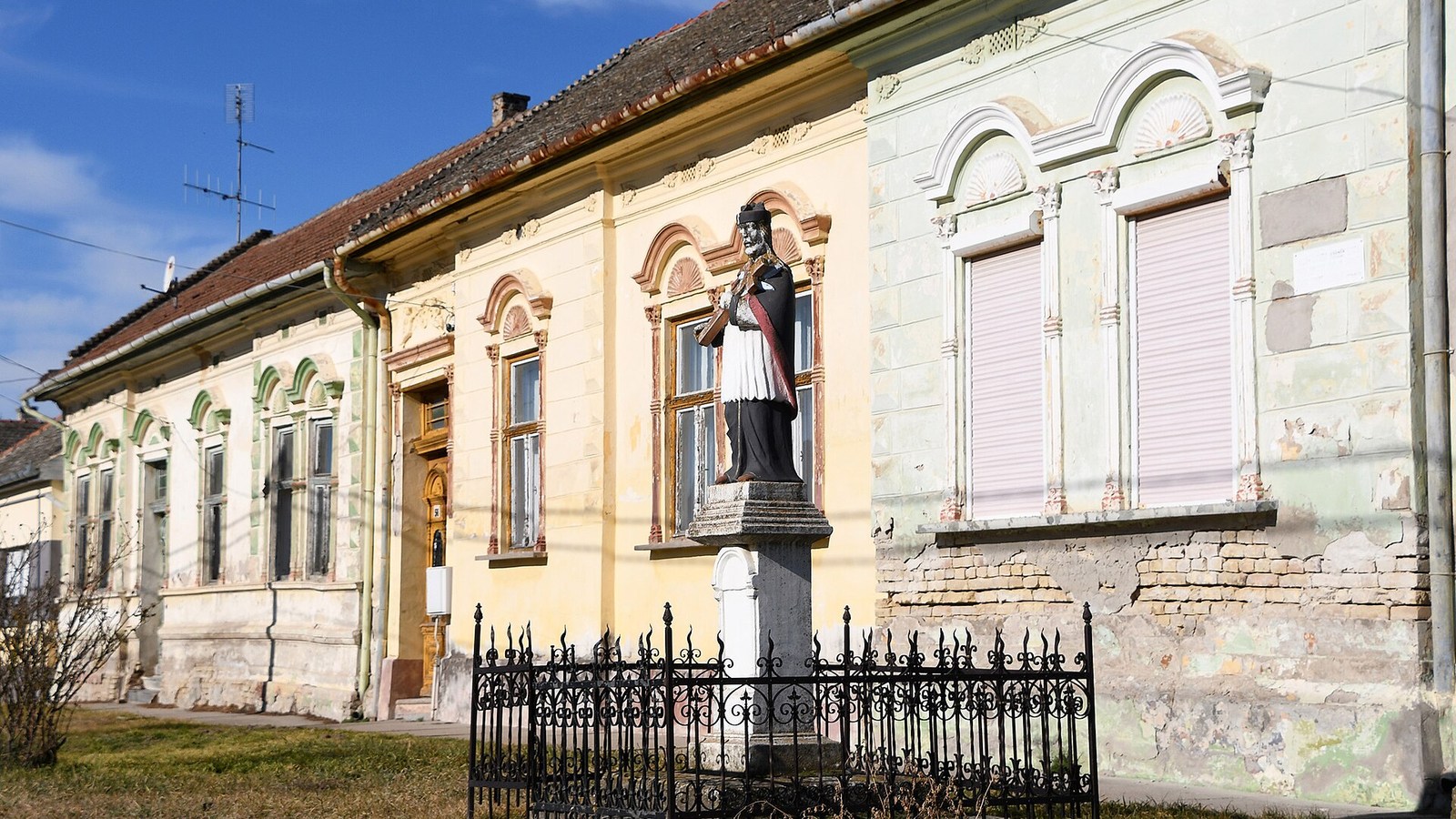 Katymár, a főutcai Nepomuki Szent János-szobor kovácsoltvas kerítéssel, mögötte díszes homlokzatú régi házak sora, kék ég alatt.