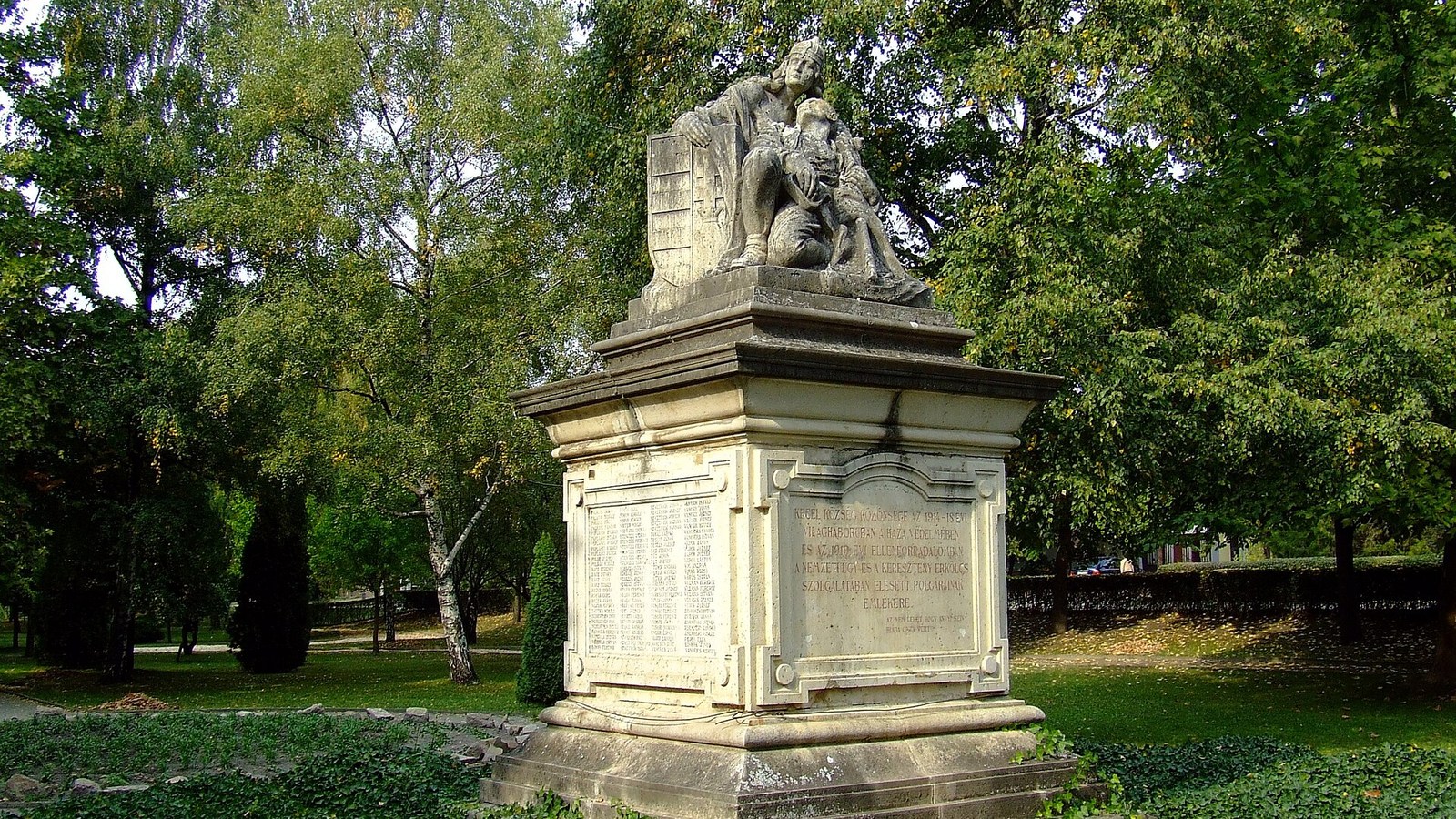 Kecel, az első világháborús hősi emlékmű sisakos katonaalakkal és sebesült társával a feliratos kőtalapzaton, a park fái között.