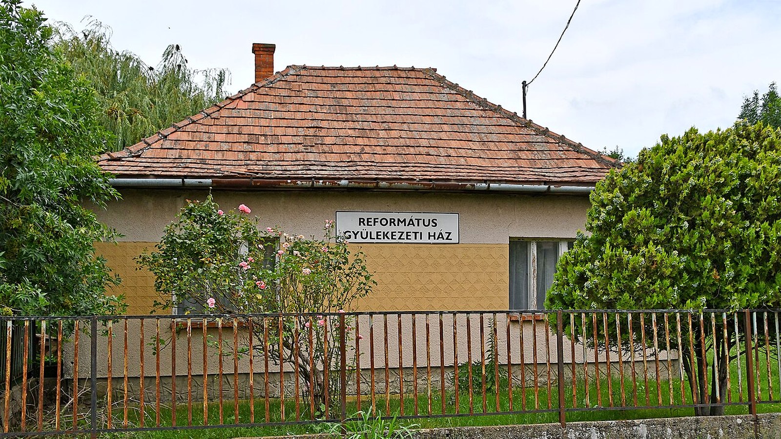 Kertészsziget, a református gyülekezeti ház cseréptetős épülete, előtte kerítés, virágzó bokrok és fák.