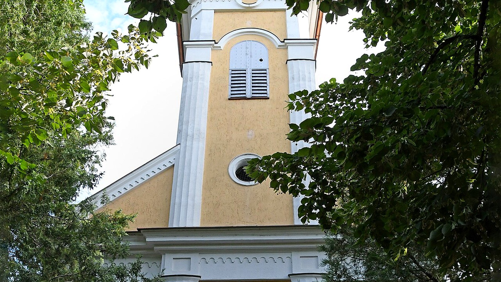Kondoros, a római katolikus templom fehér-sárga tornya lombos fák között.