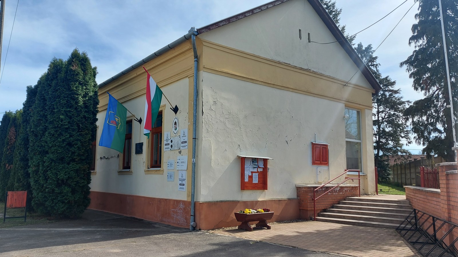 Körösnagyharsány, az önkormányzat sárga falú, nyeregtetős épülete a Kossuth téren magyar zászlóval, téglalépcsővel, virágládával és tujasorral.