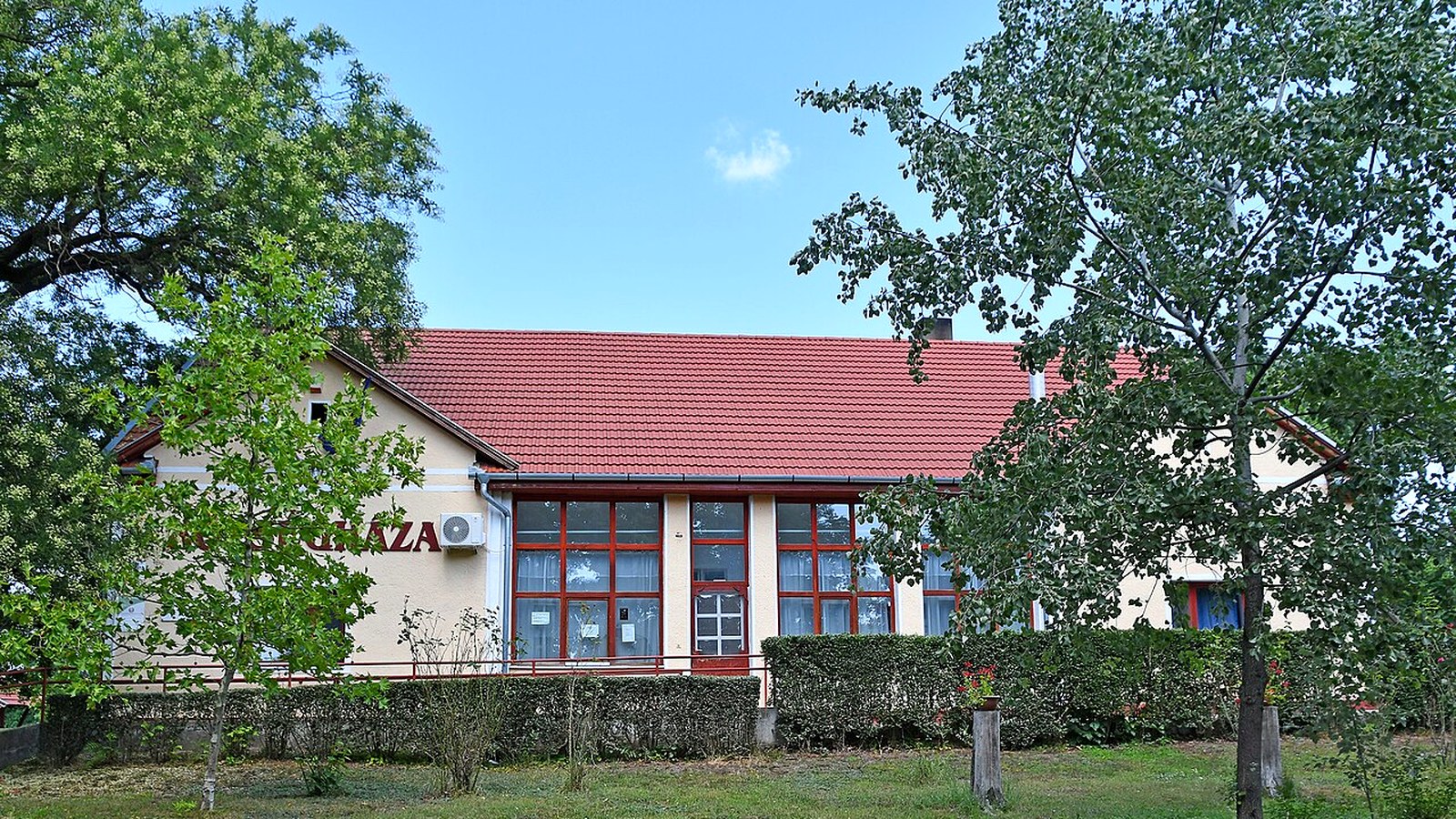 Körösújfalu, piros cseréptetős, sárga falú középület nagy ablakokkal, előtte sövény és fák.