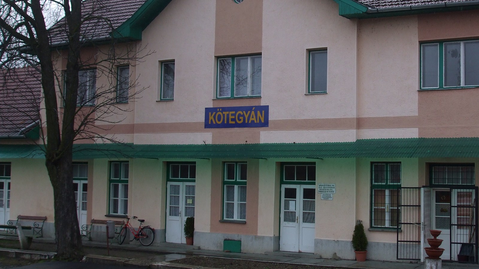 Kötegyán, a vasútállomás rózsaszín falú, zöld szegélyű felvételi épülete a Kötegyán feliratú táblával, előtte padok és egy kerékpár a peronon.