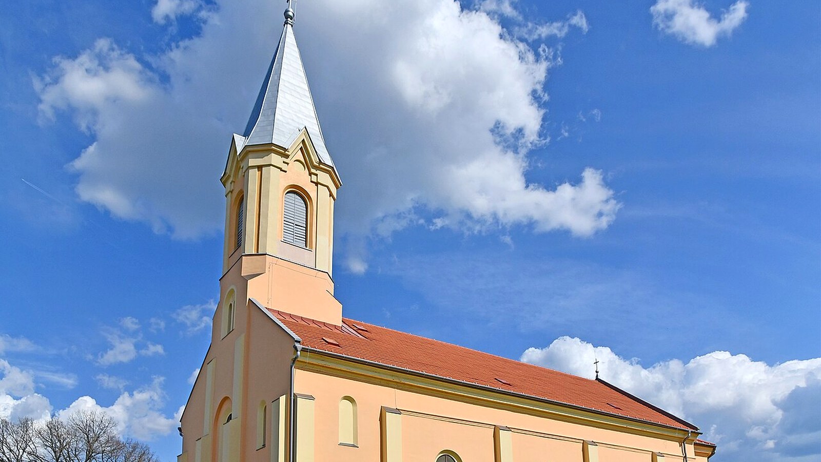 Kunágota, a templom karcsú tornya és piros tetős hajója, bárányfelhős kék ég alatt.