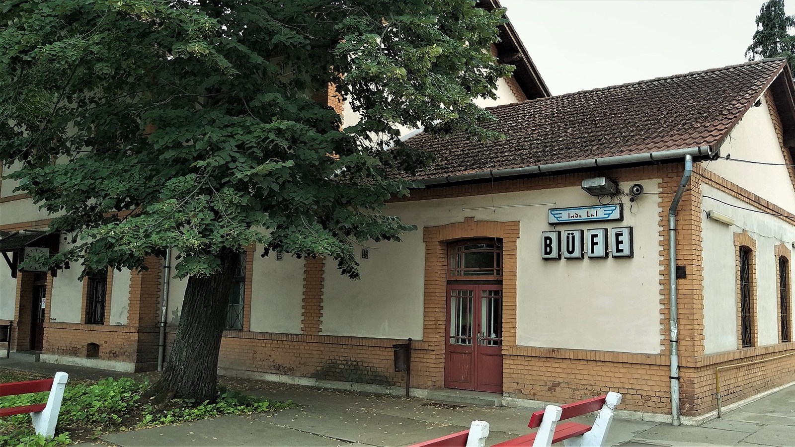 Lőkösháza, a vasútállomás téglaszegélyes, cseréptetős épületszárnya büfé felirattal és egy nagy lombos fával, előtte piros-fehér padok a peron mellett.