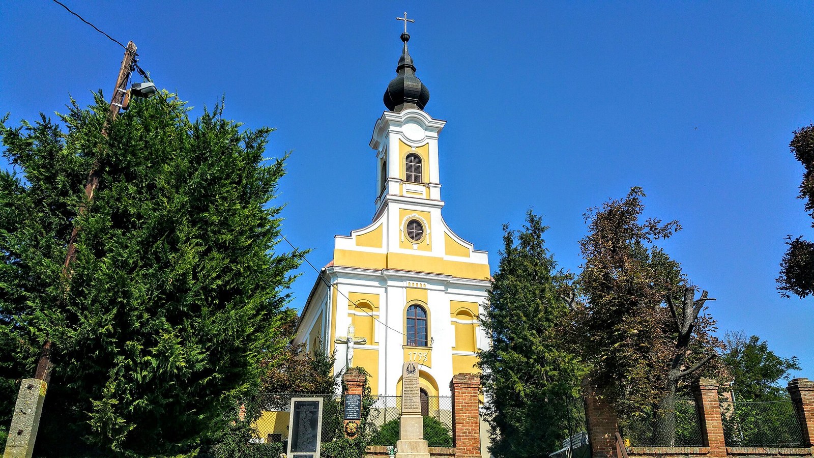 Magyarsarlós, a sárga-fehér római katolikus templom tornya a fák között, kék ég alatt.
