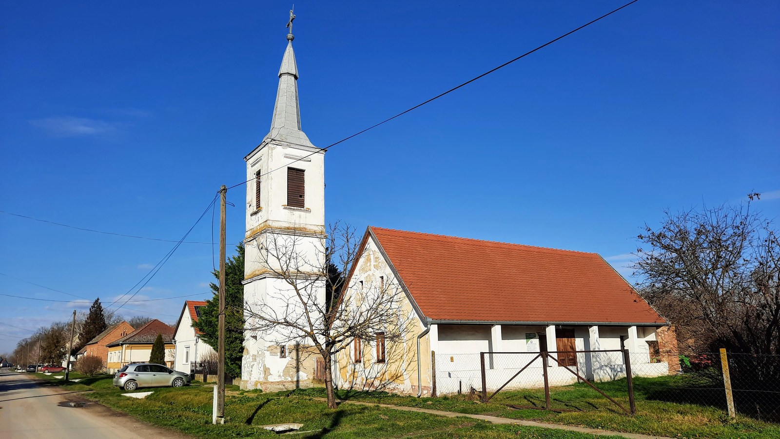 Magyartelek, a Fő utcai fehér harangtorony és mellette egy piros tetős épület, előtte füves utcaszél és falusi utca.