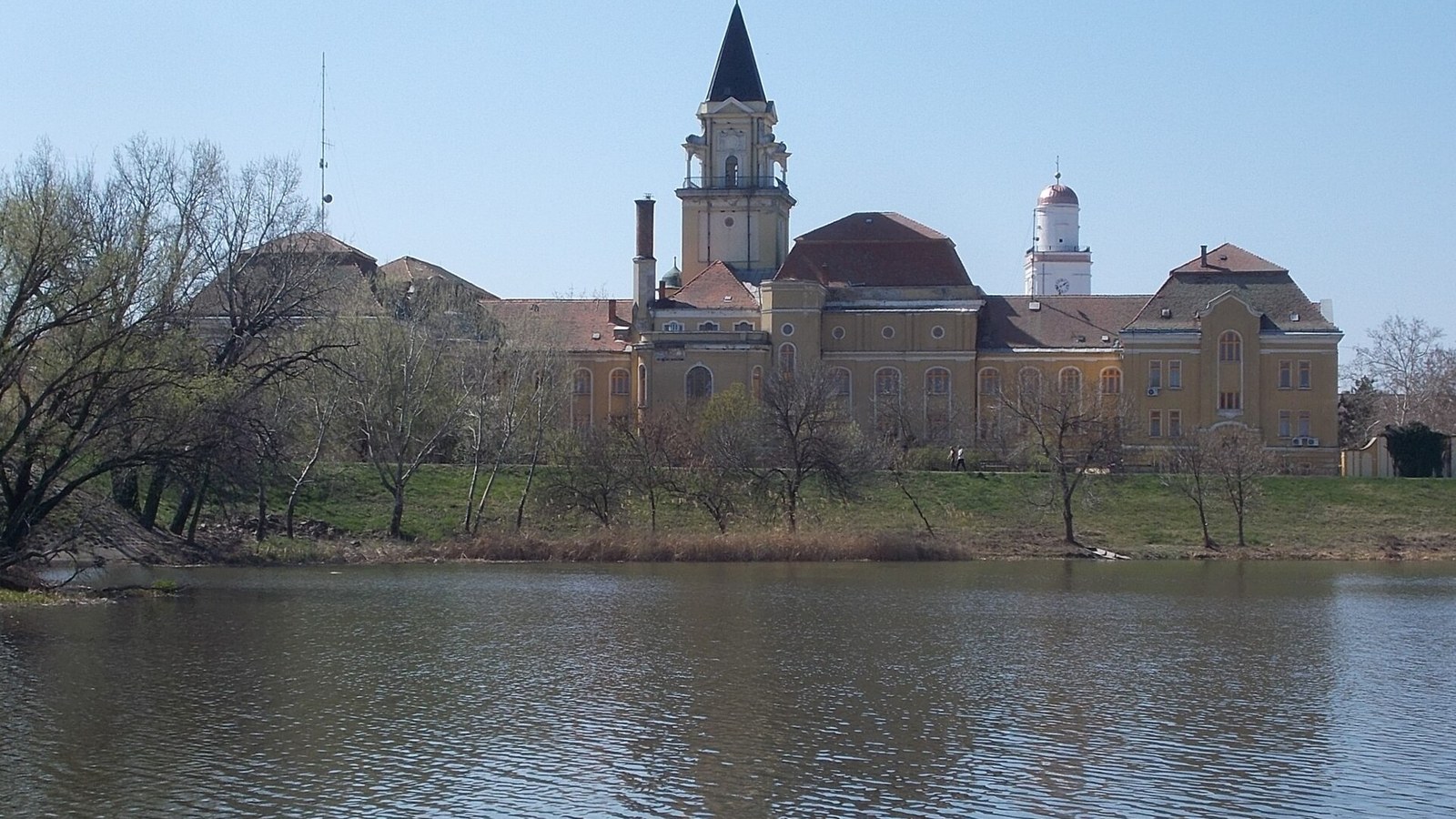 Mezőtúr, a tornyos városháza a Hortobágy-Berettyó túlpartjáról, a folyó vizével és a parti fákkal.