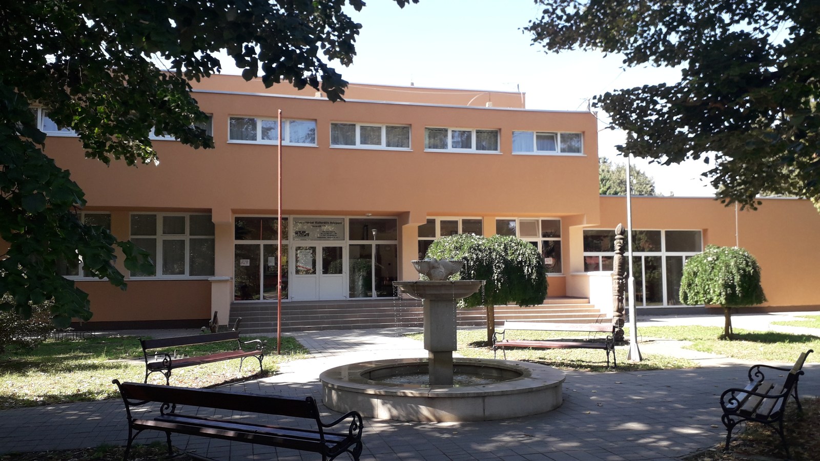 Nagyszénás, a Czabán Samu Művelődési Ház és Könyvtár narancssárga épülete előtti tér szökőkúttal, padokkal és lombos fákkal.