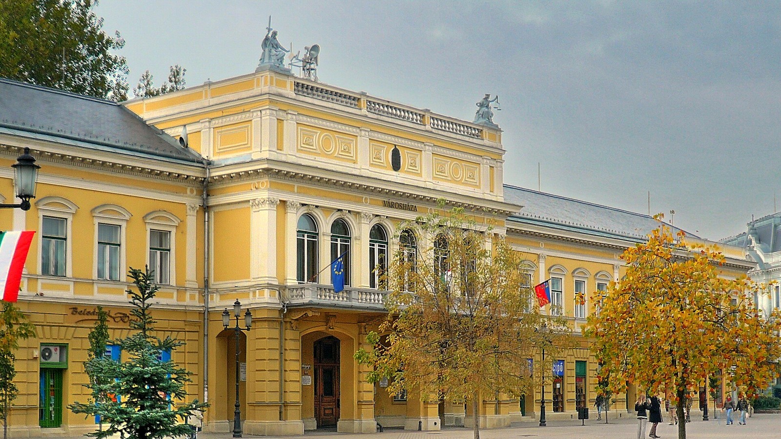 Nyíregyháza, a Városháza sárga, szoborcsoportokkal díszített homlokzata a belvárosi sétálóutcában, őszi lombú fákkal.