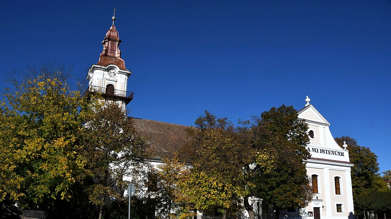 Orosháza, fehér templom vörös toronysisakkal és egy fehér oromzatos épület, őszi fák és mélykék ég.