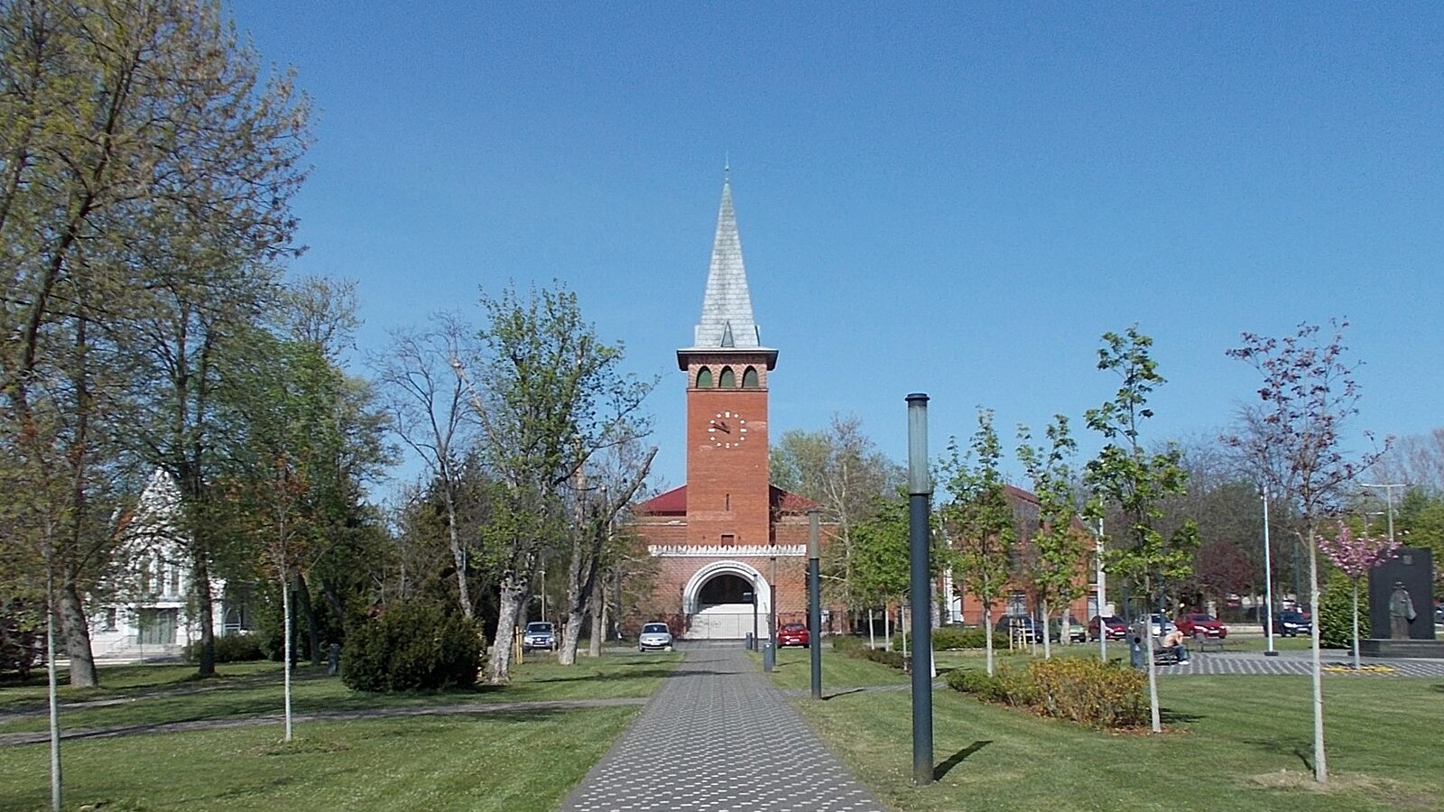 Pestszentlőrinc, a Kossuth tér parkja térköves sétánnyal és fiatal fákkal, a végén a vöröstéglás, órás tornyú református templommal.