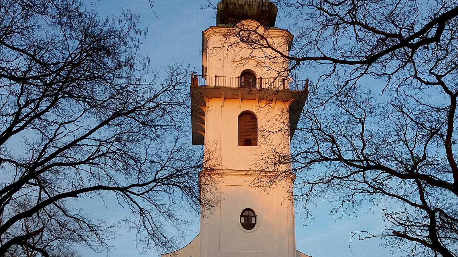 Szeghalom, fehér templomtorony kopasz faágak között, kék ég alatt.