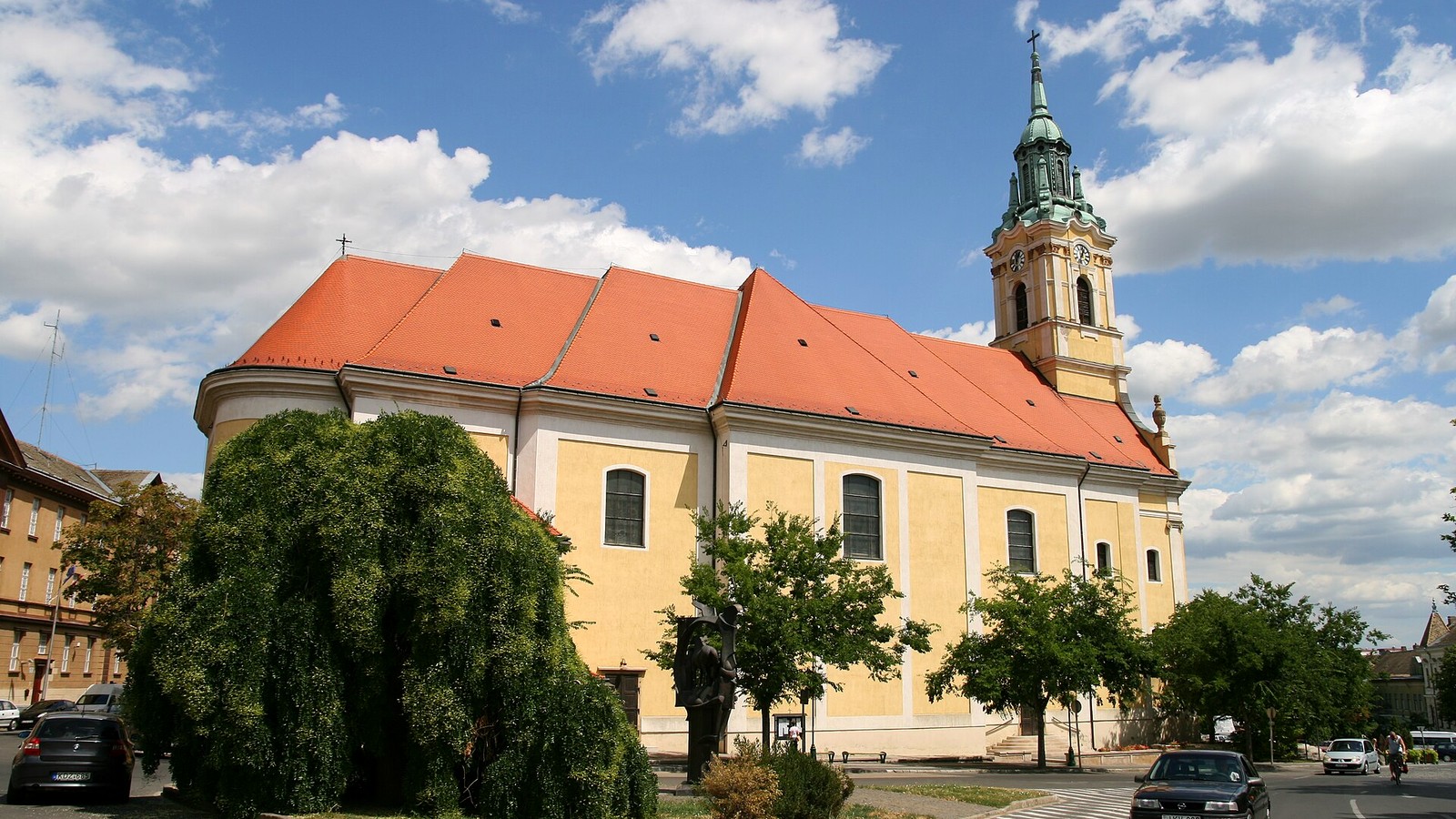 Szekszárd, a Béla király téri copf stílusú belvárosi templom narancsszínű tetővel és zöld sisakos tornyával, bárányfelhős nyári ég alatt.
