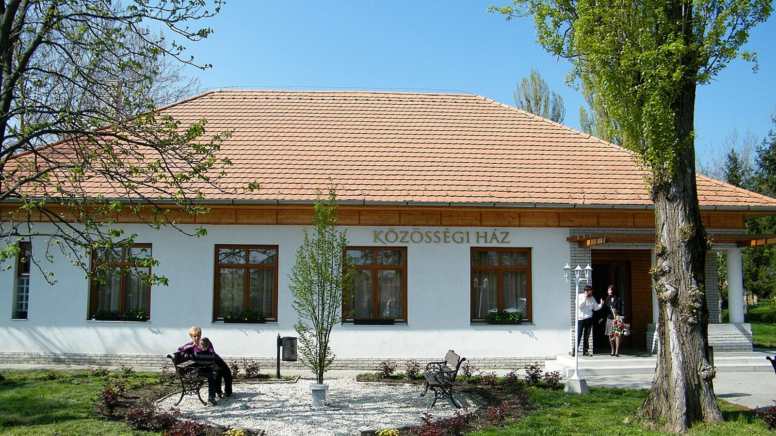 Tarhos, a Közösségi Ház fehér, cseréptetős épülete, előtte padok, virágágyás és fák.