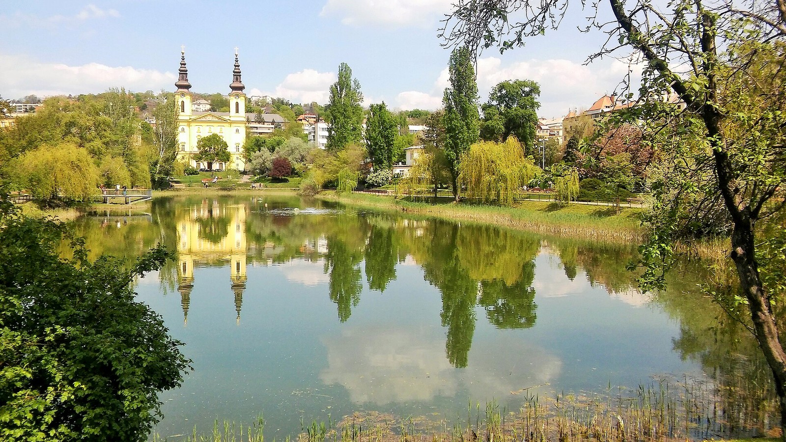 Újbuda, a Feneketlen-tó zöldellő partja szomorúfüzekkel, a háttérben a ciszterci Szent Imre-templom két tornyával.