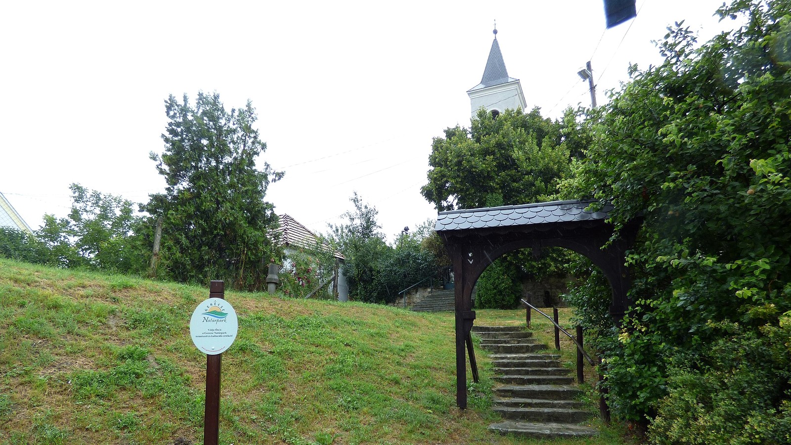 Úny, a református templom füves dombja a feljáró lépcsővel és tájékoztató táblákkal, háttérben a templomtoronnyal.