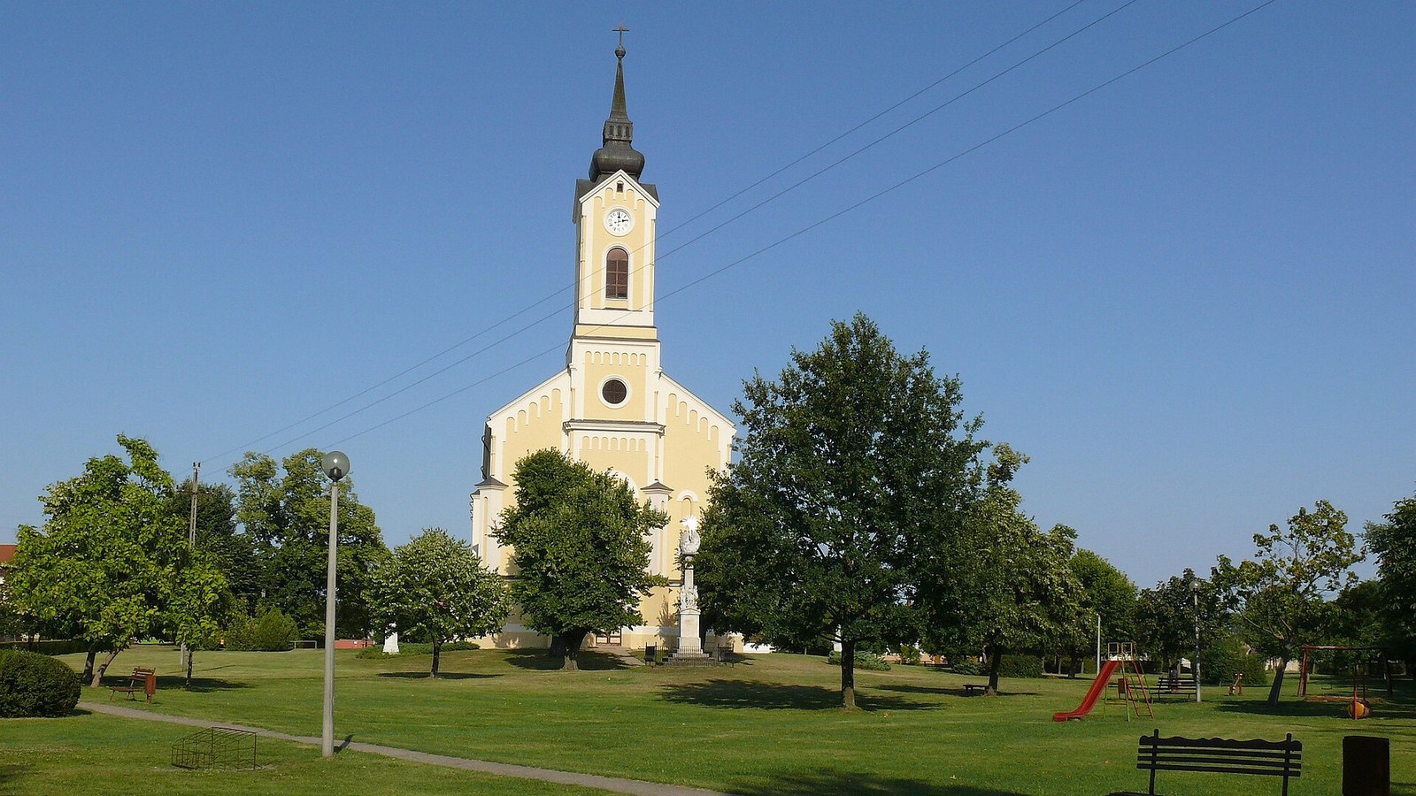 Vaskút, a Szentháromság-templom homlokzata a templomkertből nézve, előtte a Szentháromság-szoborral.