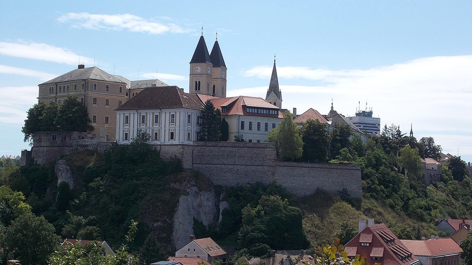 Veszprém, a Várhegy meredek sziklái fölött a várfal, a vár épületei és a Szent Mihály-székesegyház két tornya, nyári ég alatt.